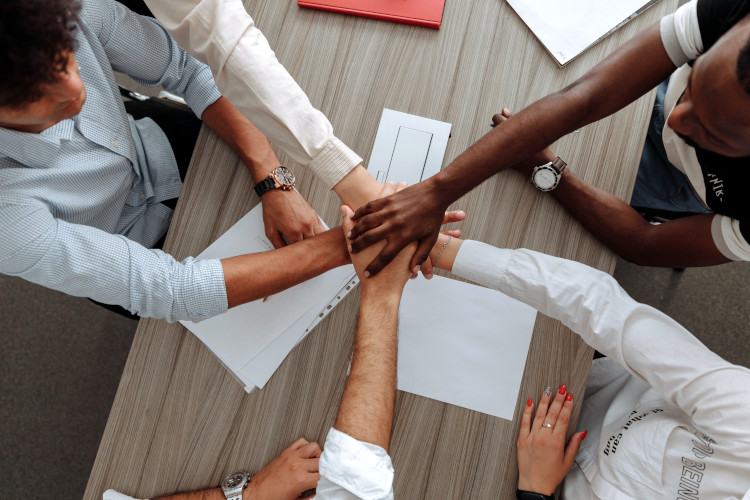 Vier Personen sitzen um einen Tisch und halten ihre Hände gemeinsam in der Mitte, symbolisierend Teamarbeit und Zusammenhalt.
